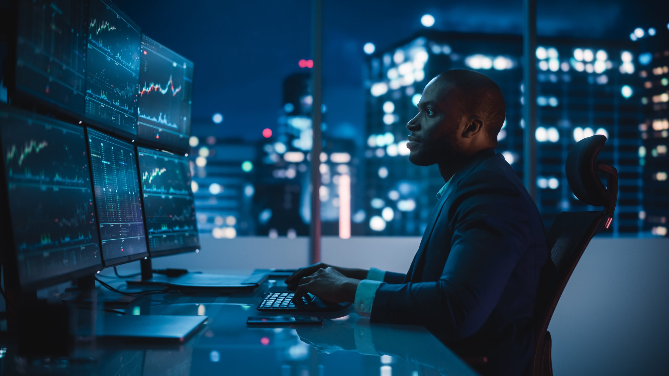 Man in a dark office monitoring multiple data screens at night, analyzing network or financial information.
