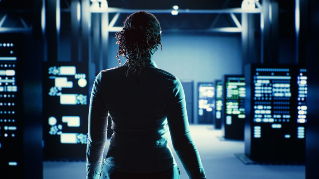 A person standing in a dark, high-tech data center, with their back to the camera. They are surrounded by rows of server racks with glowing lights, suggesting a modern and advanced security operations center.