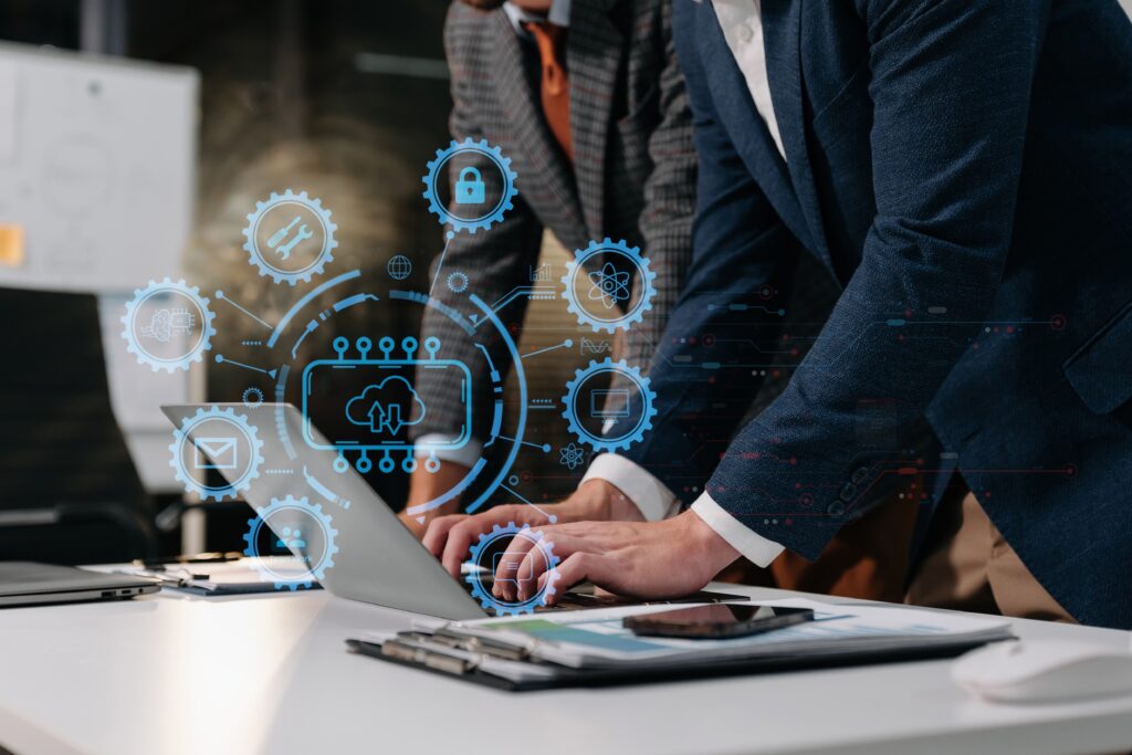 Two people in suits lean over a laptop in a modern office. Blue holographic tech icons, like clouds and gears, hover above the laptop