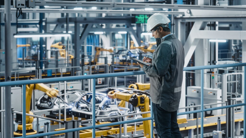 Industrial engineer using a tablet to monitor automated manufacturing equipment and robotic assembly systems inside a modern smart factory.